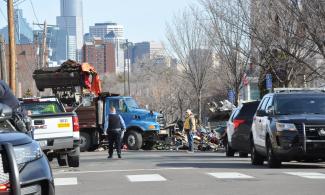 A frontloader dumps a mangled tent and wooden platform materials into a dumptruck.  A grey-mustached white man in a fluorescent vest and a police officer holding a piece of paper watches.  Several police squad cars are in the foreground.  A second frontloader is partially visible over a large pile of crushed tents, wood, bicycles, and clothing.  The Minneapolis downtown skyline is in the background.