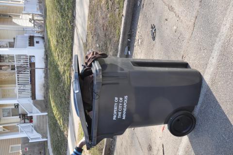 A black rectangular trash bin labeled in white block letters 'Property of the City of Minneapolis', slightly overflowing with clothing and other possessions including a brown shirt hanging out.