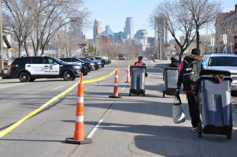 Three people, pulling wheeled municipal trash barrels behind them, walk past a line of police vehicles on a blocked-off street, with the Minneapolis skyline in the background.